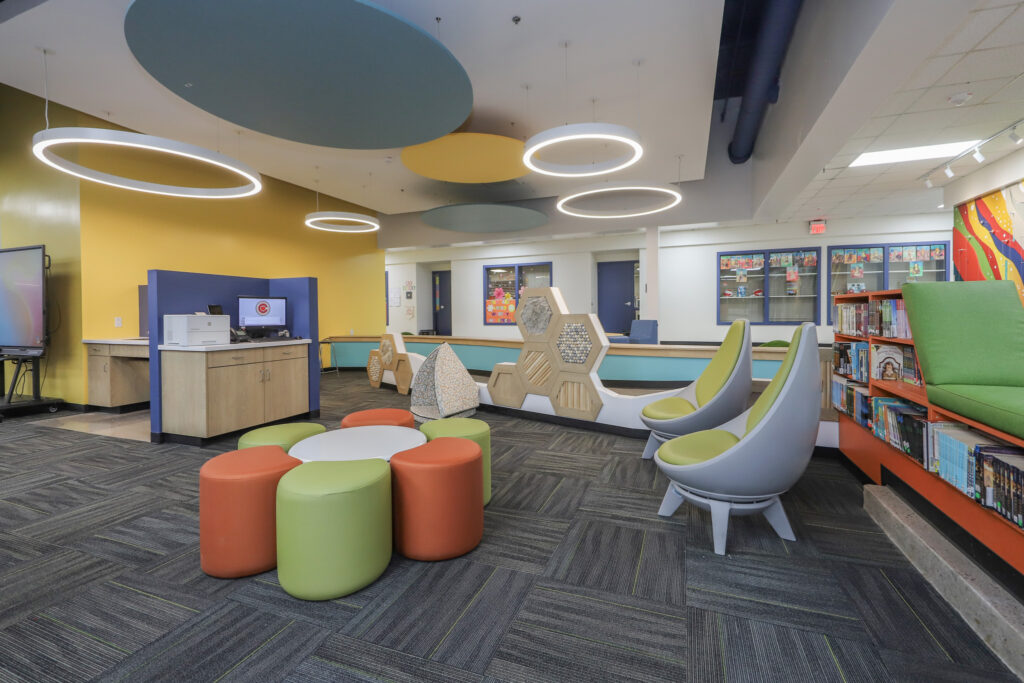 Interior of K-12 media center with orange and green stools and student furniture.