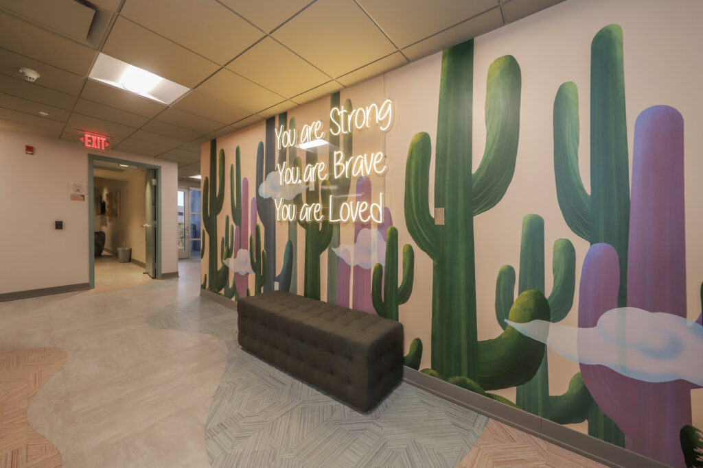 Hallway mural with saguaro cactus painted on the wall and a neon light sign that reads 'You are strong. You are brave. You are loved'